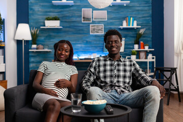 Smiling african american couple feeling happy watching TV while having a good time together sitting on sofa at home. Cheerful future parents watching family movie on television program.