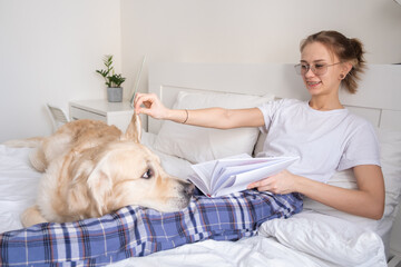 A beautiful girl is reading a book in the bedroom with her golden retriever friend. A young woman lies on the bed with her dog.