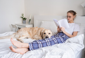 A beautiful girl is reading a book in the bedroom with her golden retriever friend. A young woman lies on the bed with her dog.