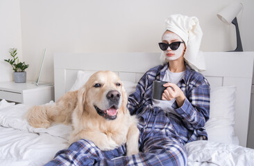 A beautiful girl does spa treatments and has fun with her dog on the bed in the morning. Young woman does a facial and strokes her dog in her bedroom