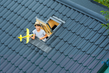 Little happy boy is playing with toy airplane in open skylight on roof house. Roof is covered with...