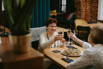 Two smiling elderly women with dark and fair hair clinking glasses of champagne sitting at table...
