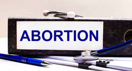 The desktop has a stethoscope, a blue pen, and a gray file folder with the text ABORTION. Medical...