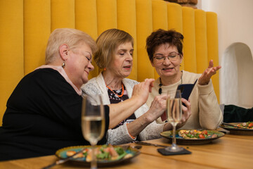 Three smiling beautiful elderly women look at mobile phone, sitting at wooden table with pizza and glasses of champagne.