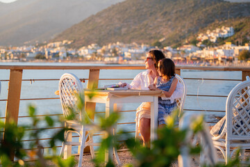 Mother with young daughter meet the morning sunrise sitting at a table outdoors by the sea. Summer holiday, rest, travel concept.