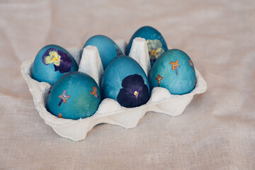 Naturally dyed cyan blue pressed flower Easter eggs in egg box on neutral beige tablecloth background. Handmade craft natural decoration.