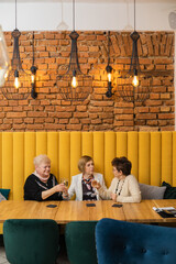 Three smiling elderly women with short dark and fair hair clink glasses of champagne sitting celebrating at restaurant.