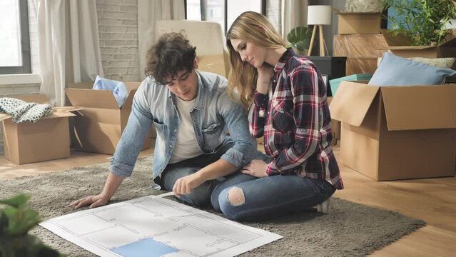 Young couple sitting on the floor looking at blueprints of the new house planning home interior on layout architectural project,moving to new apartment concept 4k