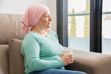 Young woman with cancer in head scarf relaxing drinking coffee sitting on a sofa in the living room in a house interior - obrazy, fototapety, plakaty