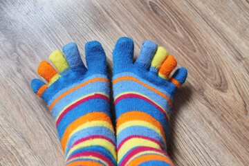 Two legs with outstretched fingers stand together on a brown laminate, close-up, top view. On the feet are socks in multi-colored stripes. Socks are worn on each finger.