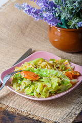 Close-up of eating cabbage fried or stewed with herbs, carrots, onions on a pink square plate, fork and knife on a tablecloth.
