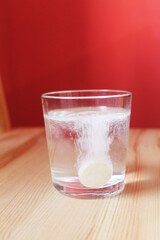 The glass is full of water with a large round tablet dissolving with bulbs. A glass stands on a brown wooden surface against a red background, close-up, side view. Poppy tablet. Vitamins. Health care.