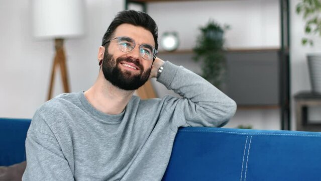 Pleasant smiling hipster guy trendy eyeglasses thinking dreaming sitting on comfortable blue couch