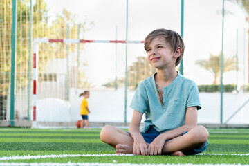 Calm boy sitting on green lawn with ball and looking seriously away © Igor
