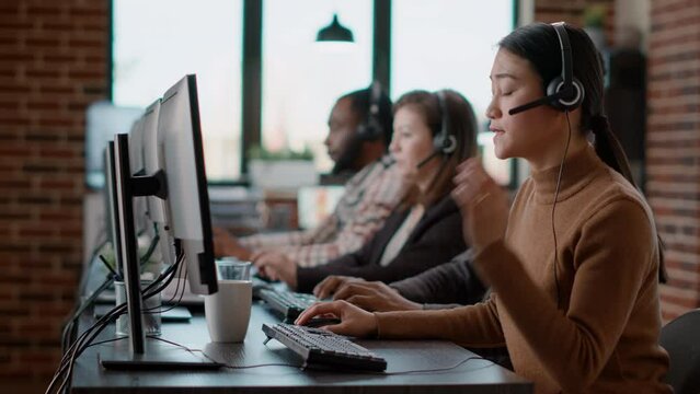 Asian woman using audio headset to have conversation at call center job with helpline clients, helping with telemarketing assistance. Customer care service employee with telesales at desk.