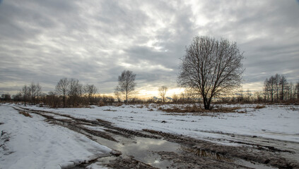 Broken dirt road with puddles in early spring. Dramatic winter landscape at sunset.