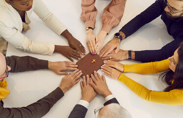 Group of several different young and senior mixed race multiracial multiethnic people who belong to the Christian church holding hands on the Holy Bible and praying together during a religious meeting