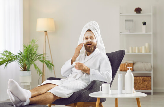 Smiling metrosexual man in bathrobe relax in chair at home spa enjoy beauty facial procedures. Happy guy take care of face skin, make treatment apply cream do mask. Skincare, cosmetology concept.