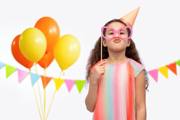 birthday, childhood and people concept - portrait of little girl in dress and party hat with glasses over balloons and flags over balloons and flags on white background