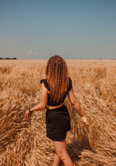 young teen girl with curly hair in black summer costume is standing from the back with hands to the side on the golden wheat field background with blue sky at sunny day. lifestyle concept, free space