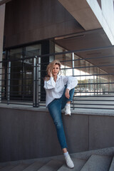 young blonde student girl in white shirt and blue jeans is sitting casual and smiling happy on the street stairs of  gray store background. lifestyle concept, free space