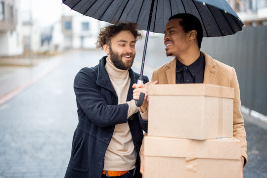 Stylish gay couple walking together with parcels on some residential street on rainy weather. Caucasian and hispanic man with umbrella wearing coats. Concept of homosexual lifestyle and delivery