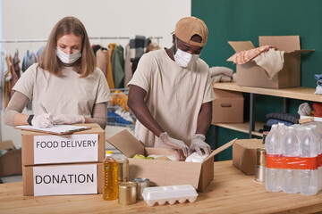 Unrecognizable man and woman wearing masks on faces standing at table packing food supplies for donation