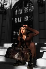 Obraz na płótnie Canvas Fashionable young brunette woman with long legs wearing solid long sleeve bodycon one piece jumpsuits posing with a leather brown travel bag