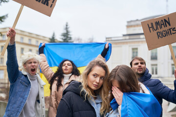 Two young caucasian women embracing in front and group of people manifesting against war in Ukraine...