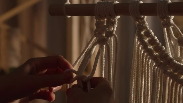 Woman weaves pattern of macrame threads with her own hands for decoration or home decor. Woman's hands tying knots on ropes in a creative workshop in soft light. Handmade. Close up. Slow motion.