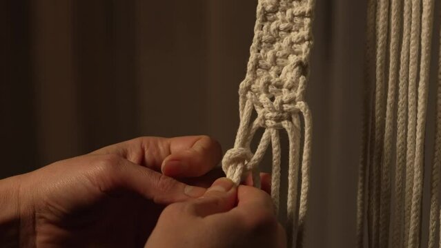 A woman makes a rope craft in a creative workshop. Woman's hands tie knots on macrame threads and create a creative lace ornament. Weaving macrame pattern in Boho style. Close up. Slow motion.