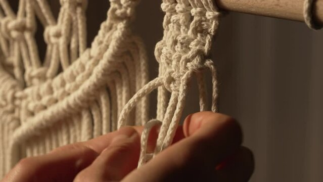 A woman makes a rope craft in a creative workshop. Woman's hands tie knots on macrame threads and create a creative lace ornament. Weaving macrame pattern in Boho style. Close up. Slow motion.