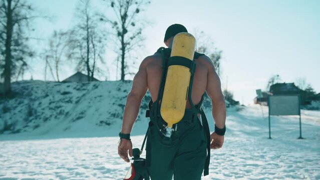 A muscular man with a naked torso with a drager on his back goes towards a snow-covered hill against the backdrop of the sun. Back view