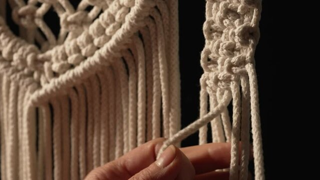Woman weaves pattern of macrame threads with her own hands for decoration or home decor. Woman's hands tying knots on ropes in a creative workshop on black background. Handmade. Close up. Slow motion.