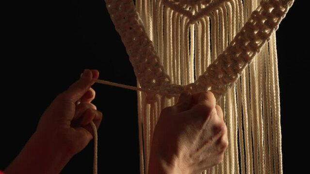 Women's hands weave a macrame pattern in creative workshop on black background. Woman ties knots on cotton threads to create lace. The base knots of macrame. Craft of rope. Close up. Slow motion.