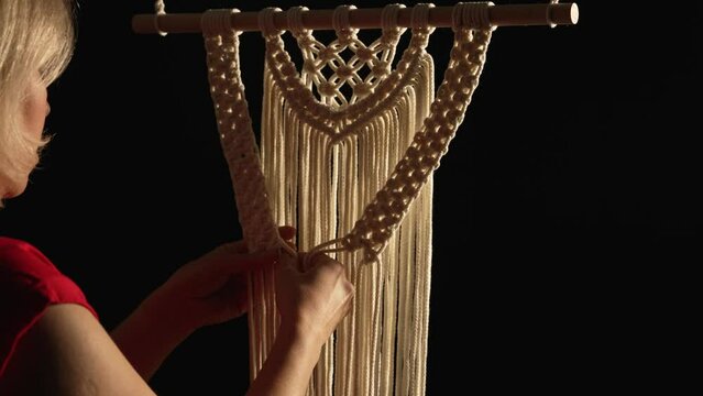 Women's hands weave a macrame pattern in creative workshop on black background. Woman ties knots on cotton threads to create lace. The base knots of macrame. Craft of rope. Close up. Slow motion.
