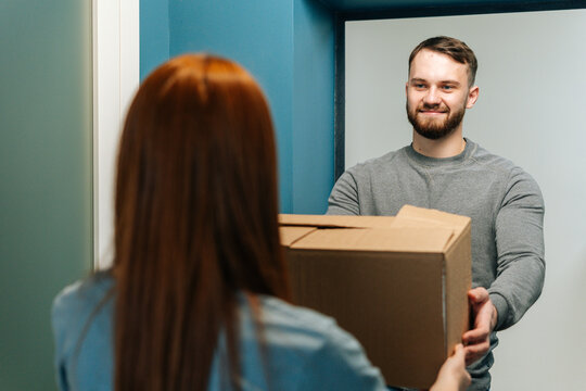 Handsome bearded courier male delivering cardboard box parcel to unrecognizable female customer on doorway at home. Rear view of young woman client receiving parcel package post from delivery man.