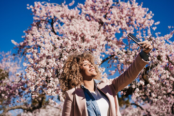 smiling hispanic woman in park taking picture with mobile phone. Spring flowers background