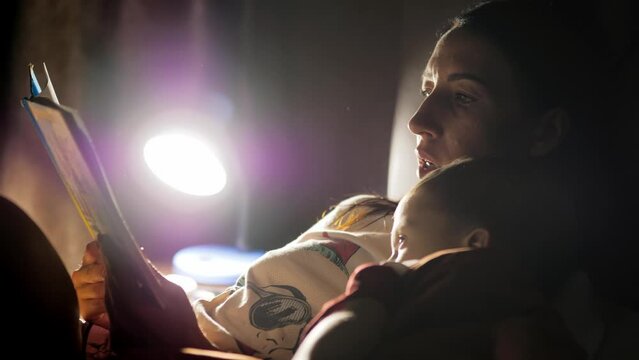 Child reading book in bed. Mother reading a bedtime story to her little son in bedroom. Education for children. Family before going to bed mother reads to her son book near a lamp in the evening.