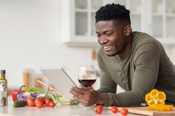 Fototapeta na wymiar Happy Black Man Using Digital Tablet And Drinking Red Wine In Kitchen