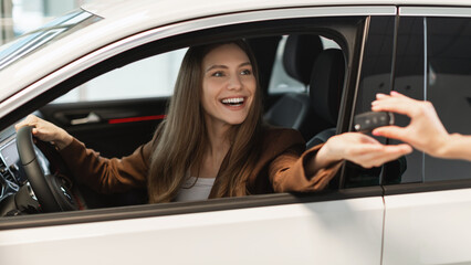 Fototapeta premium Happy young woman sitting inside modern auto, taking car key from salesperson, purchasing new vehicle at dealership