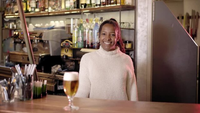 Happy smiling waitress serving a customers glass beer at the bar counter. Hospitality professional pouring an alcoholic beverage while looking at the camera.