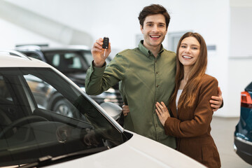 Fototapeta na wymiar Joyful young Caucasian couple holding new car key, smiling at camera in modern dealership store