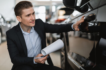 Handsome auto insurance agent sitting near new car, making checkup, taking notes at automobile dealership center