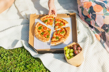 Two women eating fresh pizza from a paper box sitting on a grass on a sunny day having picnic having grapes