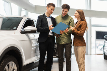Confident car salesman showing purchase details to young Caucasian couple at dealership center