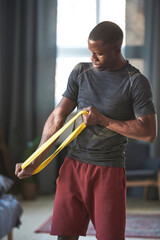 Vertical medium shot of handsome young Black man doing strengthening exercise with resistance band at home