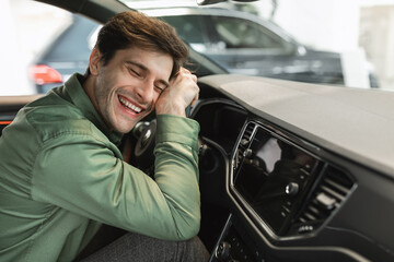 Delighted young man sitting in driver's seat, hugging steering wheel of his new car, closing eyes...
