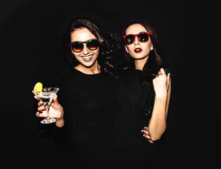 Two beautiful women black night fashion dress posing isolated on a black background. Pretty brunette girl friends twins having fun drinking cocktails. Singing and dancing. Bottle glass of alcohol.