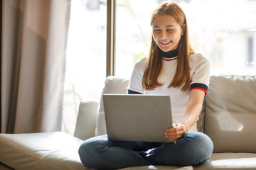 Naklejka premium Happy smiling Asian woman sitting on couch and using laptop at living room at home, watching funny video, learning language, video calling, mother working online. High quality photo
